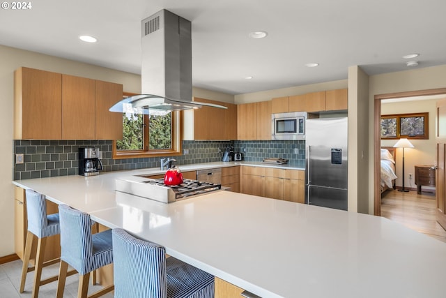 kitchen with kitchen peninsula, appliances with stainless steel finishes, backsplash, a kitchen bar, and island range hood