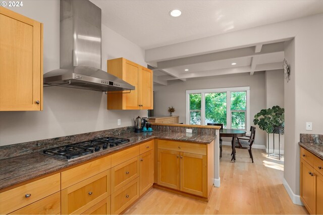 kitchen with stainless steel gas cooktop, light hardwood / wood-style flooring, kitchen peninsula, dark stone counters, and wall chimney range hood