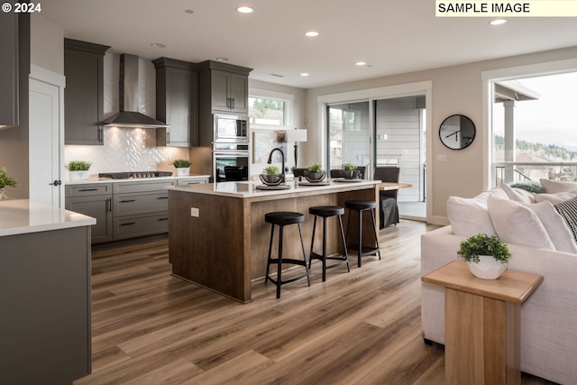 kitchen featuring dark hardwood / wood-style floors, an island with sink, wall chimney range hood, gray cabinetry, and appliances with stainless steel finishes