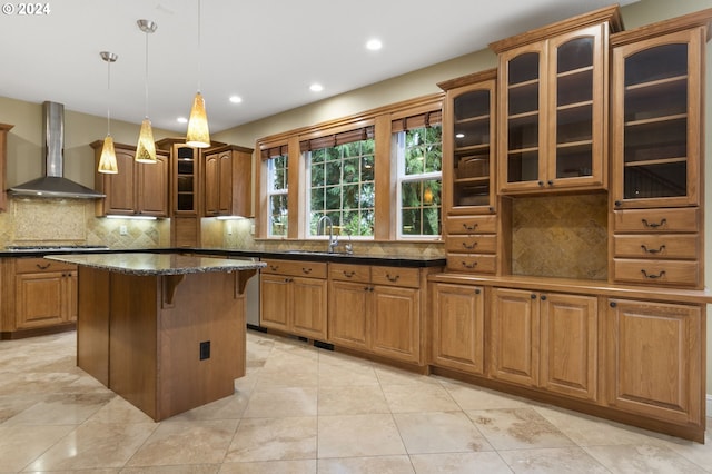 kitchen with sink, dark stone countertops, hanging light fixtures, a kitchen island, and wall chimney range hood