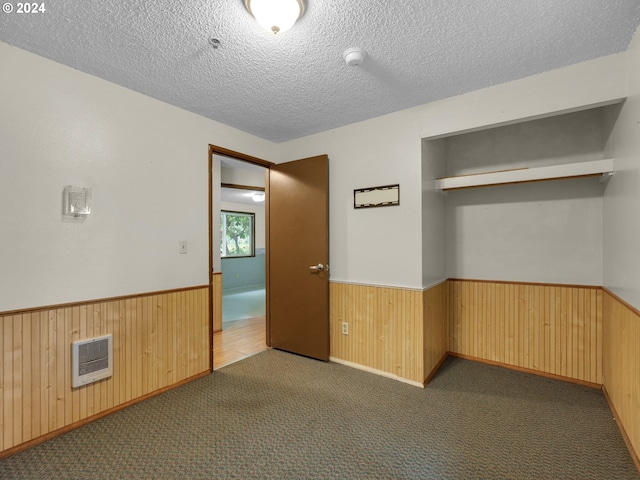 unfurnished room with a textured ceiling, wooden walls, dark colored carpet, and heating unit