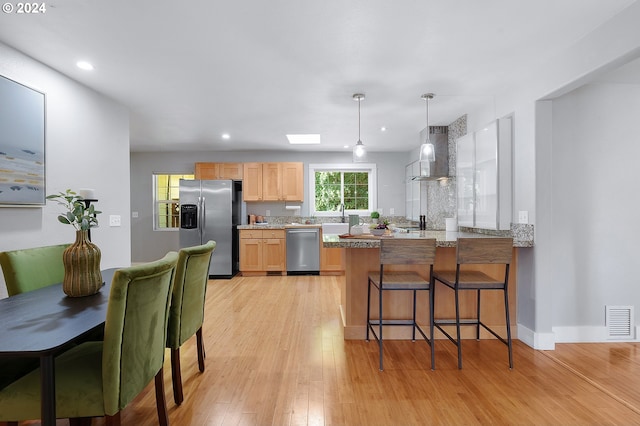 kitchen featuring light wood-type flooring, light brown cabinets, wall chimney exhaust hood, kitchen peninsula, and appliances with stainless steel finishes