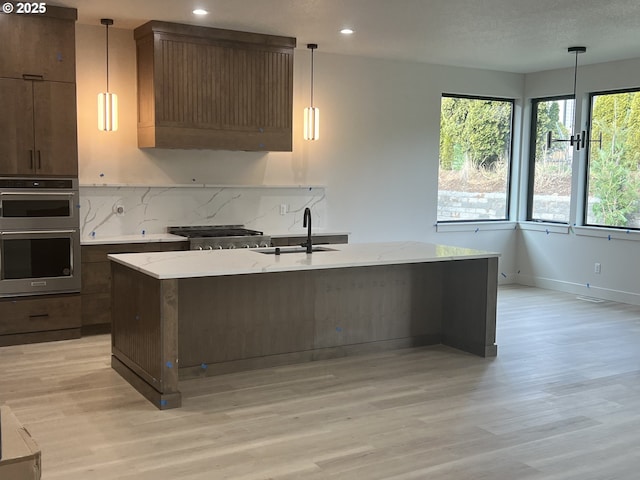 kitchen featuring sink, light stone counters, decorative light fixtures, double oven, and a kitchen island with sink