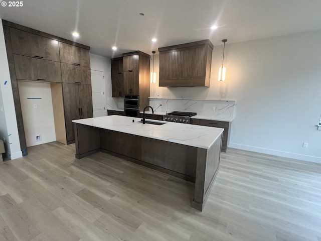 kitchen with sink, hanging light fixtures, light wood-type flooring, an island with sink, and light stone countertops