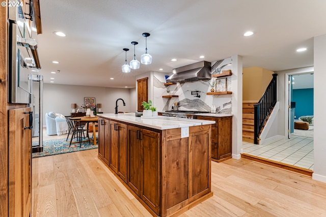 kitchen with light wood-style flooring, a kitchen island with sink, decorative backsplash, wall chimney range hood, and brown cabinets