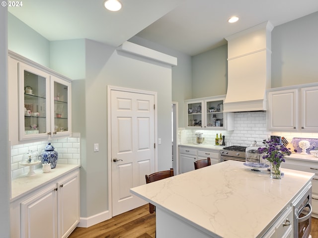 kitchen with premium range hood, light stone counters, light hardwood / wood-style floors, decorative backsplash, and white cabinets