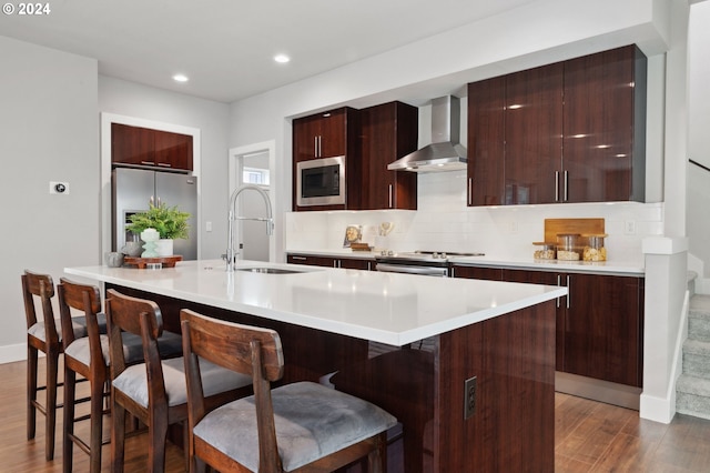 kitchen with wall chimney range hood, a center island with sink, sink, dark hardwood / wood-style floors, and appliances with stainless steel finishes