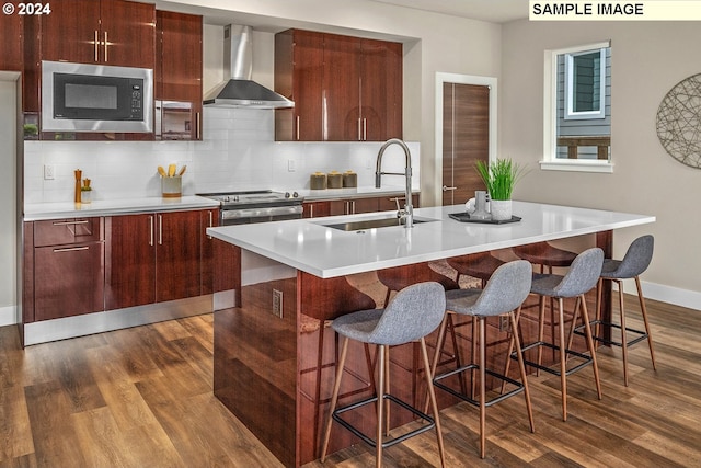 kitchen with dark hardwood / wood-style flooring, sink, a kitchen island with sink, wall chimney range hood, and appliances with stainless steel finishes