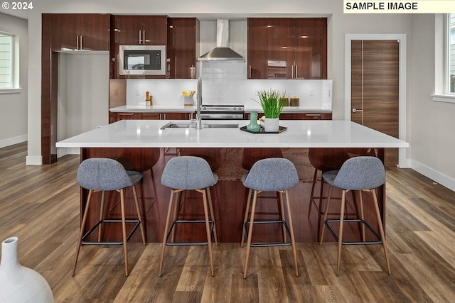 kitchen with stainless steel appliances, tasteful backsplash, a kitchen island with sink, dark wood-type flooring, and wall chimney range hood