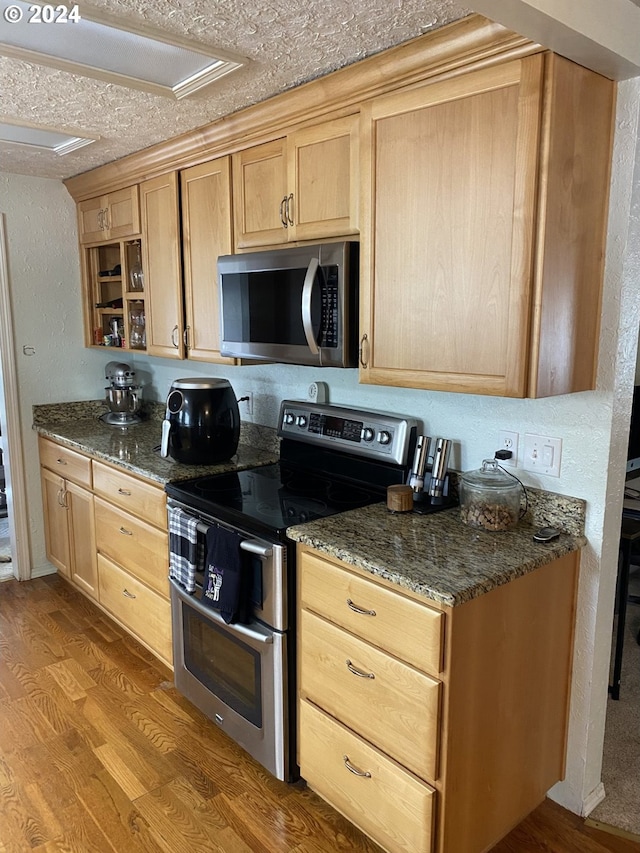 kitchen featuring light brown cabinetry, dark stone countertops, appliances with stainless steel finishes, hardwood / wood-style flooring, and a textured ceiling