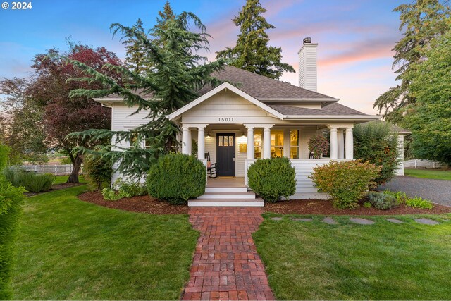 view of front facade featuring covered porch, a shingled roof, a chimney, and a lawn