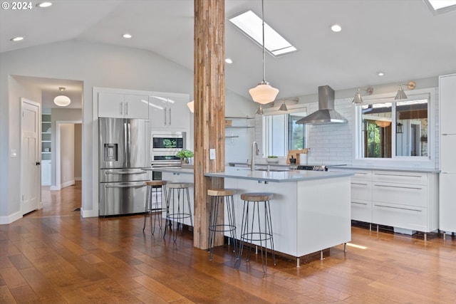 kitchen featuring stainless steel appliances, wall chimney range hood, wood-type flooring, decorative light fixtures, and white cabinets