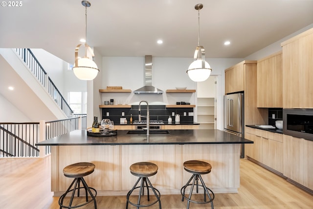 kitchen with appliances with stainless steel finishes, hanging light fixtures, light hardwood / wood-style floors, a kitchen bar, and wall chimney exhaust hood