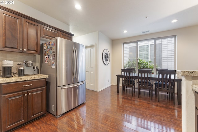 kitchen with stainless steel refrigerator, tasteful backsplash, dark wood-type flooring, dark brown cabinets, and light stone countertops