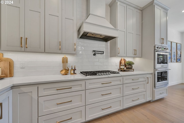 kitchen with decorative backsplash, light wood-type flooring, premium range hood, multiple ovens, and stainless steel gas cooktop