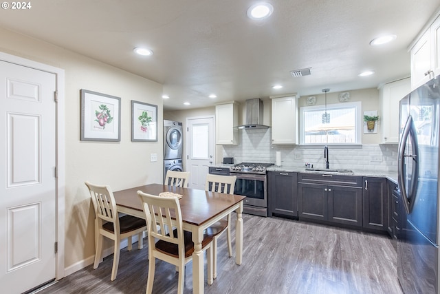 kitchen featuring a sink, wall chimney exhaust hood, light wood-type flooring, stacked washer and clothes dryer, and stainless steel appliances