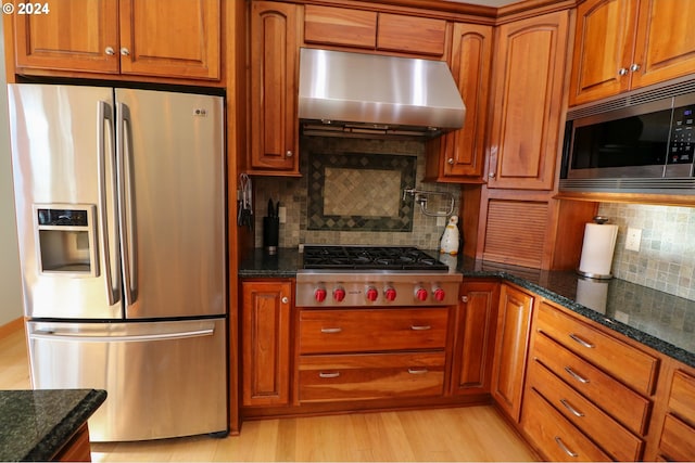 kitchen with light hardwood / wood-style flooring, dark stone countertops, stainless steel appliances, extractor fan, and decorative backsplash