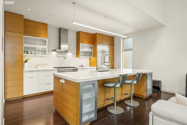 kitchen featuring backsplash, wall chimney exhaust hood, a center island with sink, stainless steel built in refrigerator, and dark wood-type flooring