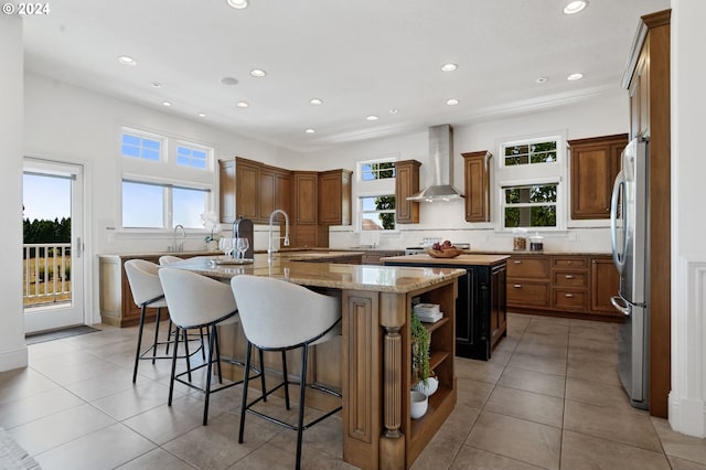 kitchen with wall chimney range hood, a center island with sink, a breakfast bar, decorative backsplash, and freestanding refrigerator