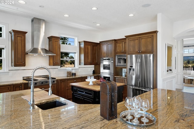 kitchen featuring a sink, light stone counters, backsplash, appliances with stainless steel finishes, and wall chimney range hood