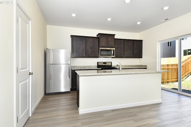 kitchen featuring appliances with stainless steel finishes, a kitchen island with sink, dark brown cabinets, light stone countertops, and light hardwood / wood-style floors