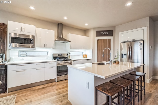 kitchen with appliances with stainless steel finishes, wall chimney exhaust hood, sink, white cabinetry, and an island with sink