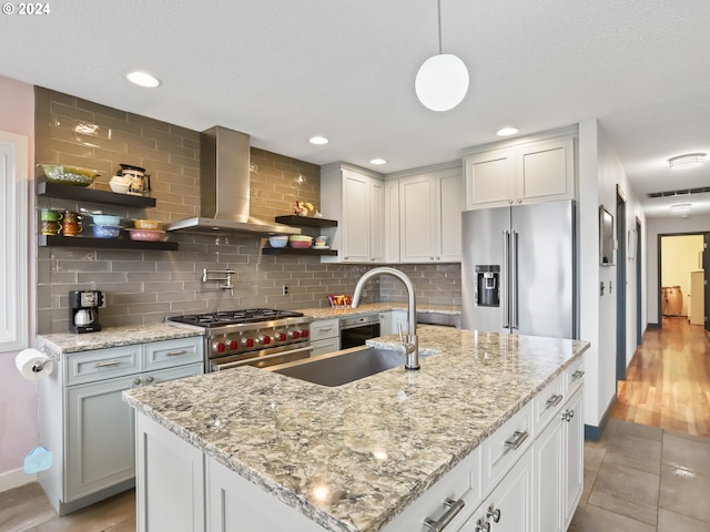 kitchen with light stone countertops, wall chimney exhaust hood, premium appliances, decorative light fixtures, and an island with sink