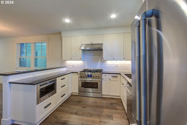 kitchen with dark wood-style floors, a peninsula, under cabinet range hood, dark countertops, and premium appliances