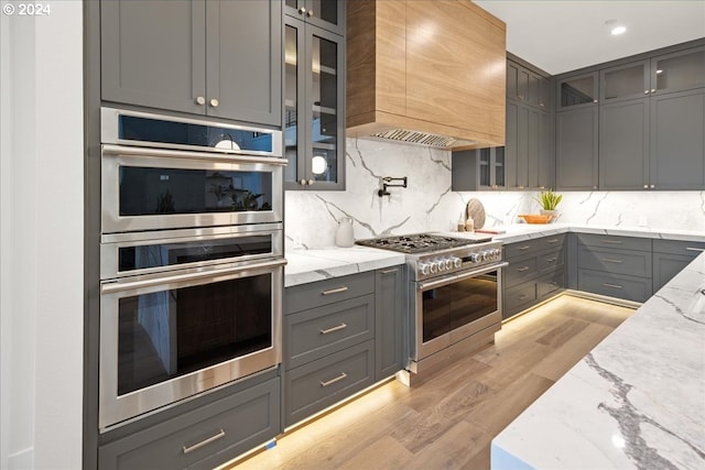 kitchen with custom exhaust hood, light stone counters, tasteful backsplash, appliances with stainless steel finishes, and light hardwood / wood-style floors