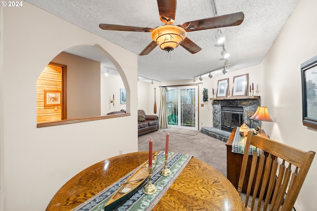 dining area featuring ceiling fan, rail lighting, a stone fireplace, a textured ceiling, and carpet