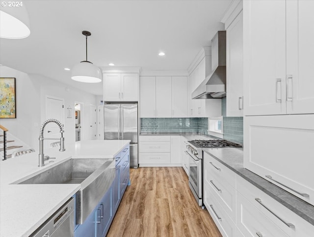 kitchen featuring wall chimney range hood, hanging light fixtures, light hardwood / wood-style floors, white cabinetry, and stainless steel appliances