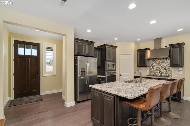 kitchen featuring dark hardwood / wood-style floors, wall chimney exhaust hood, an island with sink, sink, and appliances with stainless steel finishes