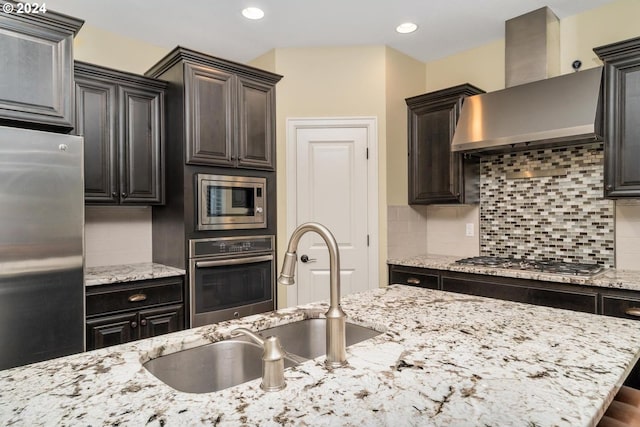 kitchen featuring light stone counters, backsplash, wall chimney exhaust hood, appliances with stainless steel finishes, and a sink
