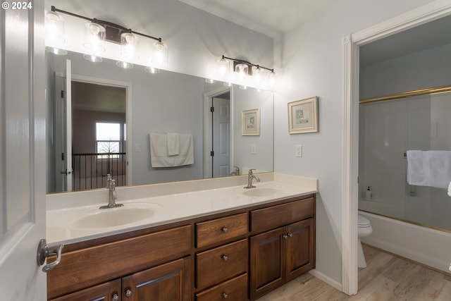 full bath featuring toilet, bath / shower combo with glass door, wood finished floors, a sink, and double vanity