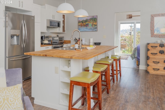 kitchen featuring appliances with stainless steel finishes, hanging light fixtures, butcher block counters, wood-type flooring, and an island with sink