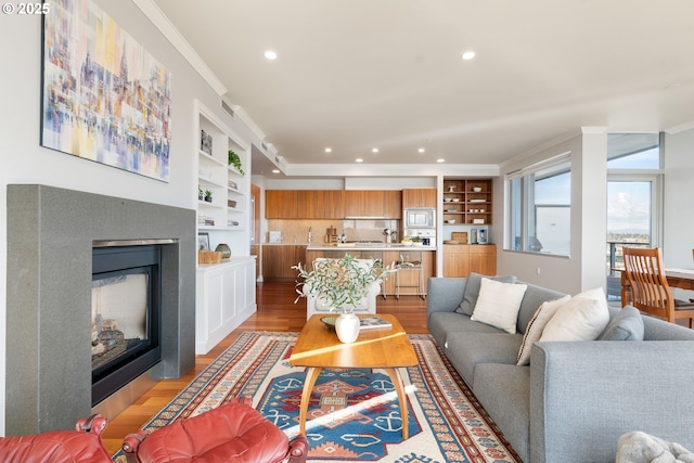 living room with built in shelves, a glass covered fireplace, light wood-style flooring, and crown molding