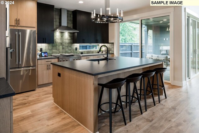 kitchen with pendant lighting, stainless steel appliances, a center island with sink, sink, and wall chimney range hood