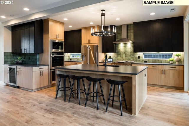 kitchen featuring wine cooler, decorative light fixtures, a kitchen island with sink, wall chimney exhaust hood, and light hardwood / wood-style floors