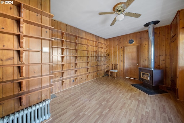 interior space with radiator heating unit, a wood stove, wood walls, ceiling fan, and light hardwood / wood-style flooring