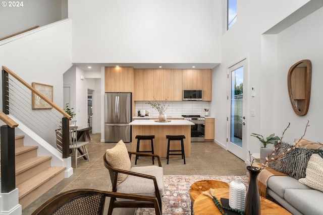 interior space featuring backsplash, appliances with stainless steel finishes, a breakfast bar, light brown cabinetry, and a high ceiling