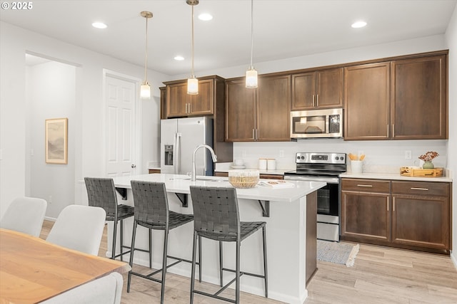 kitchen with light wood-type flooring, stainless steel appliances, a kitchen island with sink, sink, and hanging light fixtures