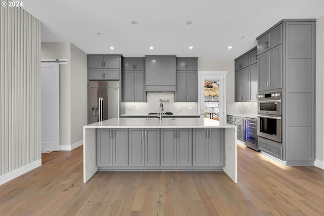 kitchen with gray cabinetry, custom range hood, stainless steel appliances, and an island with sink