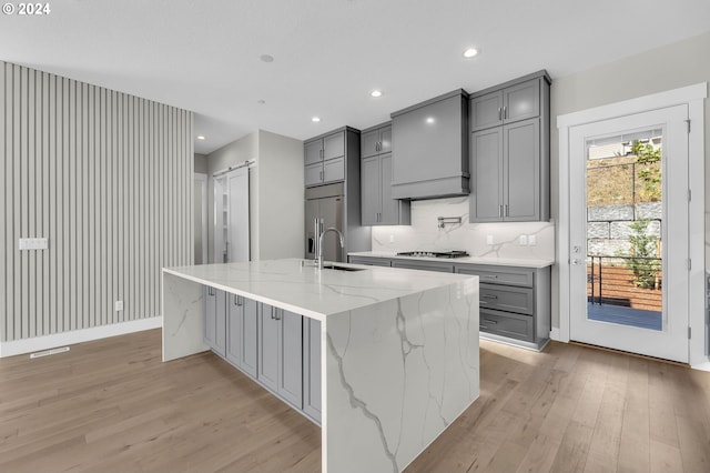 kitchen featuring gray cabinets, an island with sink, light stone countertops, custom range hood, and light hardwood / wood-style floors