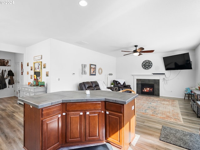 kitchen with a tiled fireplace, light wood-type flooring, a center island, ceiling fan, and tile counters