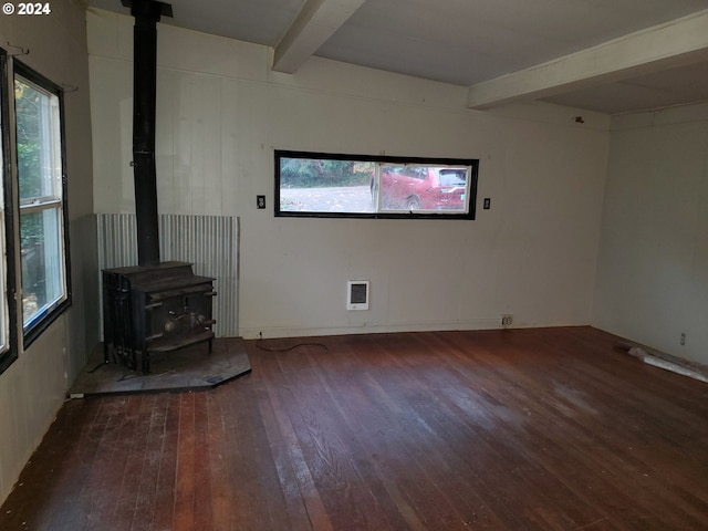 unfurnished living room featuring beamed ceiling, a wood stove, a wealth of natural light, and dark wood-type flooring