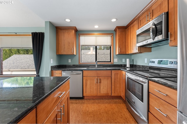 kitchen featuring a sink, appliances with stainless steel finishes, light wood-type flooring, dark stone counters, and brown cabinetry