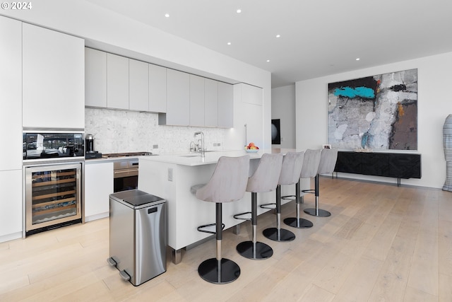 kitchen with beverage cooler, an island with sink, appliances with stainless steel finishes, white cabinetry, and light hardwood / wood-style flooring