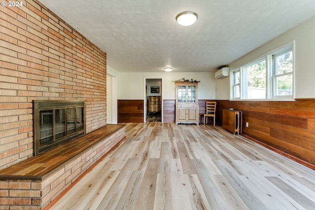 unfurnished living room featuring light wood finished floors, wooden walls, wainscoting, an AC wall unit, and a brick fireplace