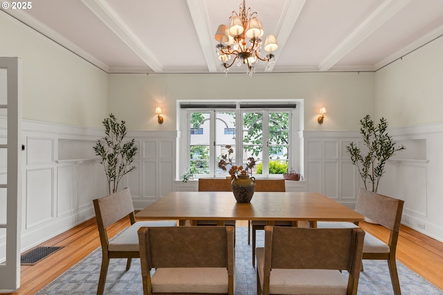 dining space with a decorative wall, a chandelier, beam ceiling, light wood finished floors, and a wainscoted wall