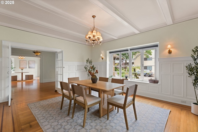 dining area with a decorative wall, a chandelier, beamed ceiling, light wood-type flooring, and a wainscoted wall